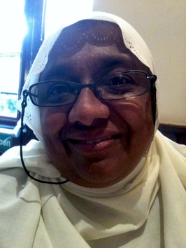 Colour photograph of the head and shoulders of a smiling Asian woman, wearing glasses and a lacy white headscarf.