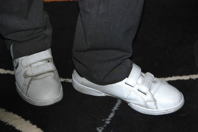 Colour photograph of a woman's feet in white trainers with velcro-based fastenings, with grey trousers above. She is standing on a black rug with white and grey patterns on it.