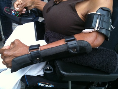 Colour photograph of a small Black woman's arm clad in a black leather arm brace as she drives her power wheelchair with the other arm.