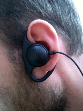 Close up colour photograph of a man's ear  with an earpiece, surrounded by dark hair and a beard.
