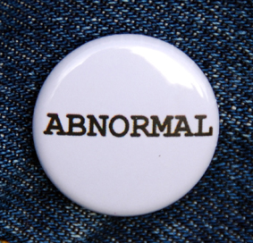 Colour photograph of a small round white badge with the word Abnormal printed in capitals in black, against a dark navy denim background