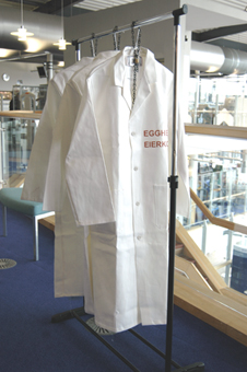 Colour photograph of three white lab coats chained to a coat rail. The coat closest to the viewer says "Egghead" on the left breast in both English and German. 