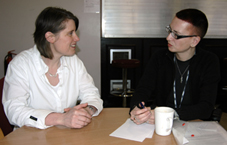 Colour photograph of Ju Gosling and Evelien sitting talking intensely at a table in the Institute canteen, with Ju making notes and drinking coffee. Evelien is a white woman in early middle age with brown collar-length hair. She is wearing a white shirt open over a white T-shirt, with a silver necklace around her neck. Ju is a white woman in early middle age with dark red hennaed hair and black-framed glasses. She is wearing a black top, and her wrists and thumbs are in black support braces.