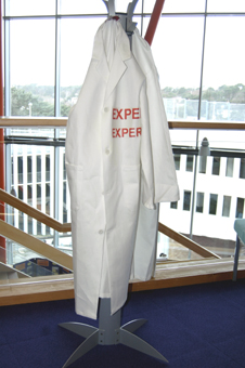 Colour photograph of two white lab coats chained to a coat rack. The coat closest to the viewer says "Expert" on the left breast in both English and German. 