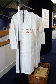 Colour photograph of three white lab coats chained to a coat rail. The coat closest to the viewer says "Genius" on the left breast in both English and German.  Next to the coats sits a laundry basket with an artist's apron inside.