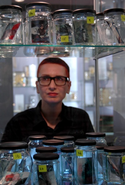 Colour photograph of the artist seen through two shelves of glass jars displayed on glass shelves. Each jar contains a photograph and has been numbered using a fluorescent sticker.