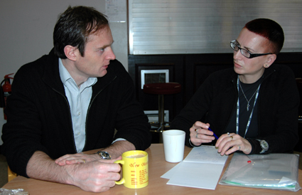 Colour photograph of Ju Gosling and Malcolm Logan sitting talking intensely 
  over coffee in the Institute canteen, with Ju making notes. Malcolm is a white 
  man in early middle age with slightly thinning brown hair. He is wearing an 
  open dark top over a pale blue shirt. Ju is a white woman in early middle age 
  with dark red hennaed hair and black-framed glasses. She is wearing a black 
  top, and her wrists and thumbs are in black support braces.
