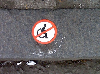 Small colour photograph of the 'Wheelchair user barred' sticker stuck on a kerb.