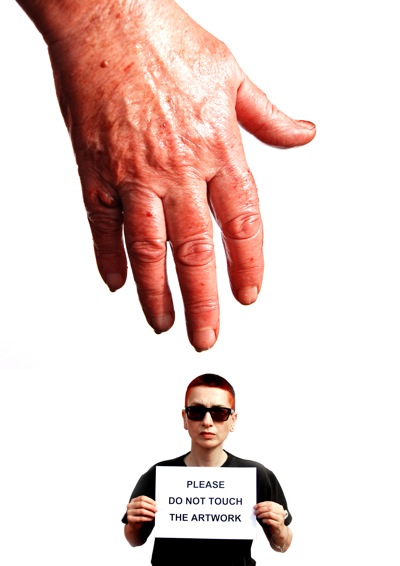 The photo shows a hand reaching down from the top of the picture, covered in blood. 
At the bottom of the picture, a woman with short red hair, dressed in a black 
t-shirt and wearing black glasses, holds up a sign saying Please do not touch 
the artwork. The hand is much bigger than the woman. 