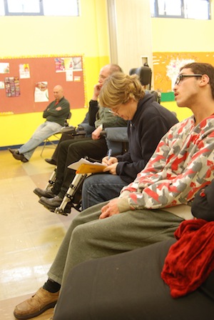 Colour photograph showing Clare Summerskill sitting writing as other workshop participants view an improvisation.